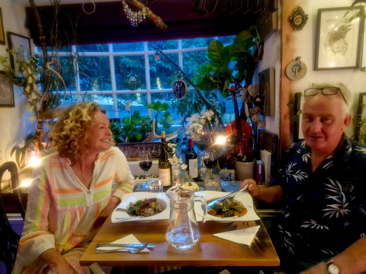 Two diners at a window table full of plants, with wine and stew
